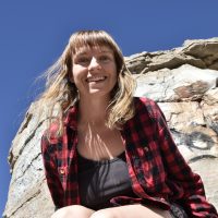 erin smiling wearing a plaid shirt on a big rock with a blue sky behind her